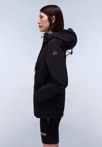 Giacca nera impermeabile con cappuccio, chiusura frontale a zip, emblema sulla manica, tasche laterali e polsini elasticizzati. Modello che indossa pantaloni corti neri.