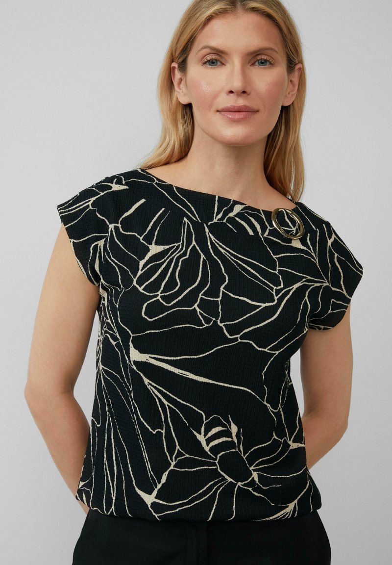Zwart kortemouwen top met een wit bloemenpatroon, textuurstof en een ronde accent bij de schouder. Ontspannen pasvorm.