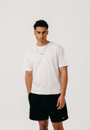 Mand med kort krøllet hår iført en hvid t-shirt, sorte shorts og en sølvkæde, står foran en ensfarvet hvid baggrund.