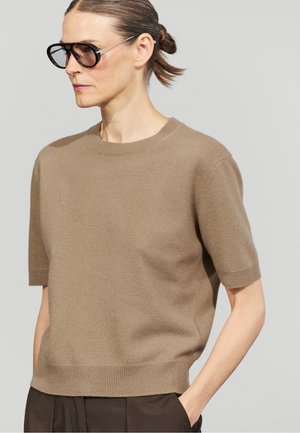 Donna con i capelli legati indossa grandi occhiali da sole scuri, un maglione taupe a maniche corte e pantaloni scuri con la mano in tasca.