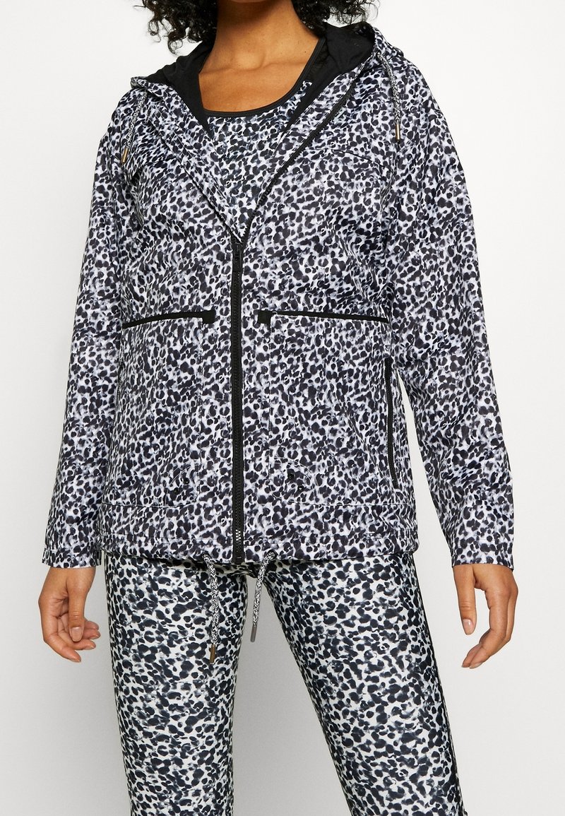 Femme portant une veste zippée à capuche imprimé léopard noir et blanc avec des leggings assortis sur un fond uni.