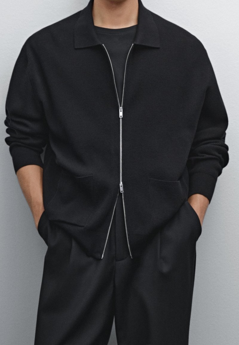 Schwarze, gerippte Strickjacke mit Reißverschluss und Kragen, ausgestattet mit zwei Fronttaschen und einem Metallreißverschluss. Kombiniert mit schwarzen, locker sitzenden Hosen.