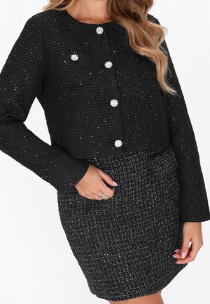 Femme portant une veste noire texturée à manches longues avec des boutons ornés et une jupe taille haute assortie.