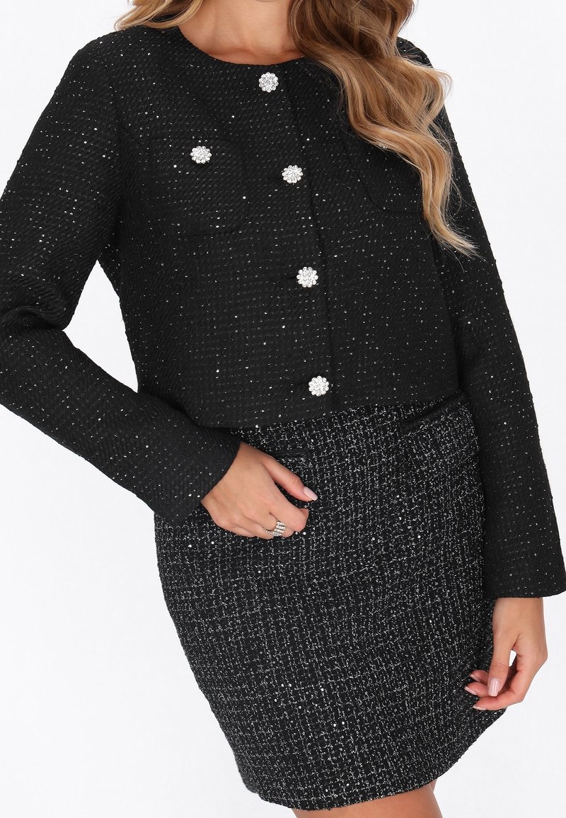 Femme portant une veste noire texturée à manches longues avec des boutons ornés et une jupe taille haute assortie.