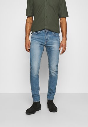 Mann trägt hellblaue Slim-Fit-Jeans, olivgrünes kurzärmeliges Hemd mit Knöpfen und schwarze Wildleder-Stiefeletten; steht vor weißem Hintergrund.