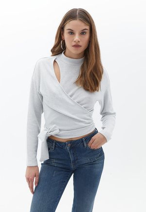 Hellgraues Wickeloberteil mit langen Ärmeln, hohem Halsausschnitt und Ausschnittdetails. Kombiniert mit dunkelblauen Jeans im klassischen Schnitt und leichter Abnutzung.