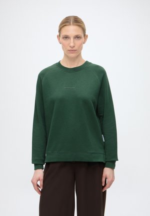 Kvinde med blondt hår iført en mørkegrøn langærmet sweatshirt og mørkebrune bukser, stående mod en ensfarvet hvid baggrund.