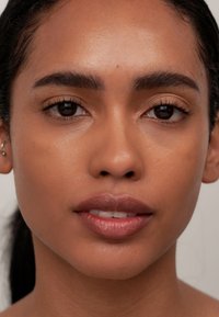 Nahaufnahme eines Gesichts mit glatter, brauner Haut, dunklen Augen, definierten Augenbrauen und vollen Lippen; das Haar ist nach hinten gegelt.