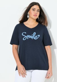 Námořnické tričko s krátkým rukávem a výstřihem do V, s bledě modrým textem "Smile". Měkká látka s volným střihem. Nosené s bílými kalhotami.