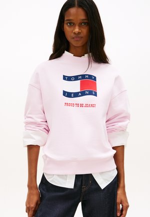 Giovane donna che indossa una felpa rosa chiaro con il logo Tommy Jeans e la scritta "Fiera di essere jeans!", abbinata a dei jeans scuri.