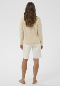 Haut beige à manches longues, tissu léger ; short blanc longueur genoux avec poches arrière ; sandales plates ; cheveux bruns ondulés.