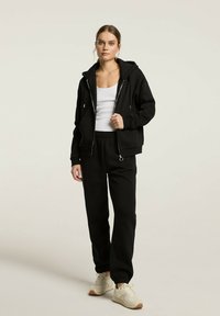 Czarna bluza z kapturem na zamek błyskawiczny z przednią kieszenią i sznurkiem, w połączeniu z czarnymi spodniami na elastycznym pasie i białymi trampkami.