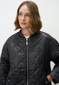 Veste bombardier noire matelassée avec un design texturé en vagues, zip argenté et poches latérales. Le matériau semble brillant et léger.