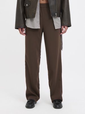 Persona che indossa pantaloni marroni larghi con vita elastica, scarpe di pelle nere, camicia a quadri non infilata sotto giacca marrone, in piedi su sfondo bianco.