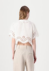 Hvit crop top med bølgekant, med utskårne blomsterdesign, korte ermer og rund krave. Sammen med lyse beige bukser.