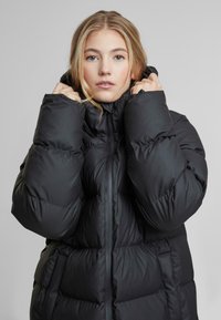 Musta puhvitakki korkealla kauluksella, jossa on tikattu design, vetoketjusuljenta ja ylisuuret hihat lisälämpöä ja mukavuutta varten.