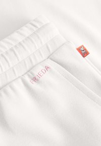 Close-up van witte elastische tailleband met roze geborduurde tekst "FRIEDA" en een klein oranje label met een witte hondenicoon.