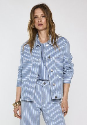 Vrouw met lang haar die een blauw-wit verticaal gestreepte jas en bijpassende broek draagt, geaccentueerd met gouden armbanden en ringen.