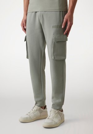 Homme portant un pantalon de survêtement cargo vert clair avec le logo « GUESS » sur la cuisse et des baskets blanches, debout devant un fond uni.