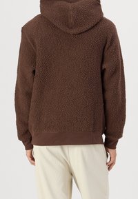 Hnědá fleecová mikina s texturovaným, chlupatým povrchem, s kapucí na stahovací šňůrku a žebrovanými manžetami, v kombinaci se světle zbarvenými kalhotami.