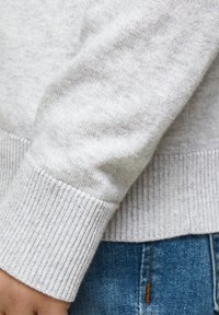 Manica di un maglione lavorato a maglia grigio chiaro con dettaglio a coste, che mette in risalto la morbida texture e il design aderente, abbinato a jeans blu.