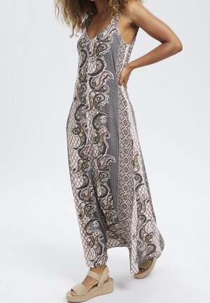 Kobieta w bezrękawnikowej sukience maxi z brązowo-czarnym wzorem paisley i geometrycznym, do tego beżowe sandały na platformie.