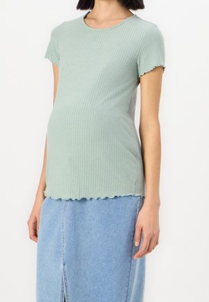 Kobieta ubrana w jasnozielony prążkowany top z krótkim rękawem i jasno-niebieską spódnicę dżinsową na białym tle.
