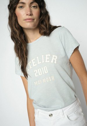 Jonge vrouw met lang bruin haar, gekleed in een lichtblauw gestreept T-shirt met de tekst "ATELIER 2010 MOS MOSH" en een witte broek, tegen een effen achtergrond.