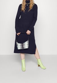 Robe à col roulé en maille bleu marine avec une fente latérale, associée à des bottines à talons en cuir verni vert néon et un sac à main rayé noir et blanc.