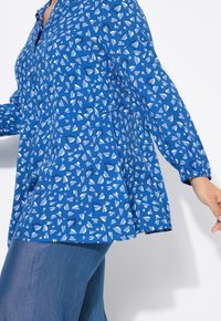 Blå blus med hjärt-mönster i ljusare nyanser, knappstängning fram, långa ärmar med elastiska muddar och en lös, svängig passform.