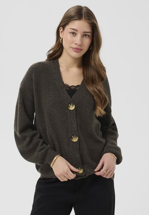 Mladá žena s dlhými hnedými vlasmi, oblečená v tmavohnedom zapínacom kardigane, čiernom topu s čipkovým lemom a zlatými šperkmi na jednoliatom pozadí.