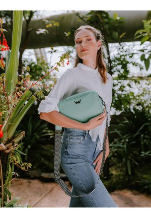 Hellmintgrüne Crossbody-Tasche mit strukturierter Oberfläche, versehen mit einem Logo und einem gemusterten Riemen in Rot, Schwarz und Weiß.