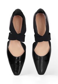 Ballerine in vernice nera con punta affilata e lati ritagliati, caratterizzate da una larga fascia elastica nera incrociata. Soletta beige visibile.