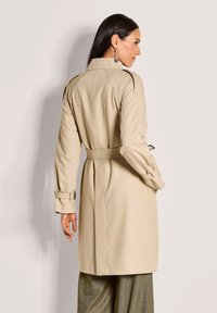 Vrouw met lang donker haar, gekleed in een beige trenchcoat met riem en grijze wijde broek, staand in zijaanzicht tegen een lichte achtergrond.