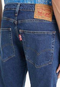Los jeans de mezclilla azul cuentan con una parte trasera alta con una etiqueta de parche de cuero marrón. Diseño de cinco bolsillos con costuras amarillas y una etiqueta de logo roja en el bolsillo trasero.