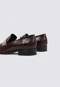 Mocasines de cuero marrón con una suela negra plana, que presentan un acabado suave y un diseño de punta redonda. Sin acentos decorativos visibles.