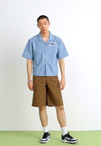 Kortermet blå skjorte med knapper og lapp, brune cargo-shorts og sorte joggesko med hvite detaljer. Enkelt design, avslappet passform.