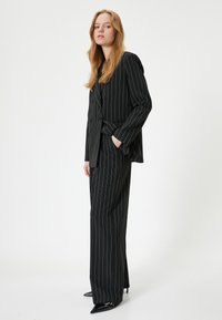 Costume noir à rayures avec une veste croisée sur mesure et un pantalon à jambes larges. Présente un tissu lisse et des chaussures noires pointues.