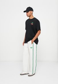 T-shirt noir avec un logo vert et rouge, associé à un pantalon large blanc avec des bandes latérales vertes et un logo brodé. Casquette noire et chaussures grises.