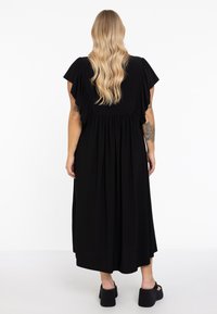 Schwarzes Kleid bis zum Knie mit kurzen, ausgestellten Ärmeln und gerafftem Bund. Glatte Textur mit fließendem Stoff. Mattes Finish, kombiniert mit schwarzen Schuhen.