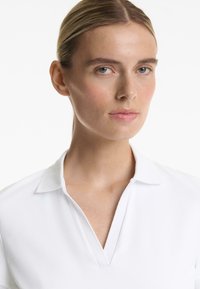 White polo shirt with a V-shaped neckline and folded collar, displayed on a neutral background. Smooth texture with a classic design.