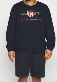 Felpa blu navy e pantaloni corti abbinati, in misto cotone. La felpa presenta un logo ricamato bianco e rosso con un design a scudo e testo.