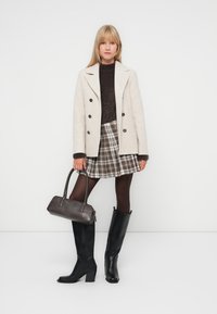 Manteau beige croisé, pull en tricot marron, jupe à carreaux, bottes noires à talons hautes jusqu'aux genoux et un sac à main sombre, sur un fond uni.