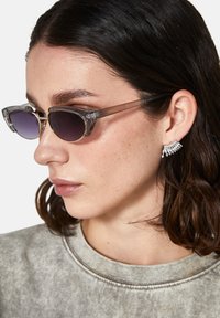 Kissakatse sunglasses, joissa on läpinäkyvät harmaat kehykset ja tummat linssit; metalliset yksityiskohdat. Yhdistetty harmaaseen sweatshirtiin ja korukorvakoruun.