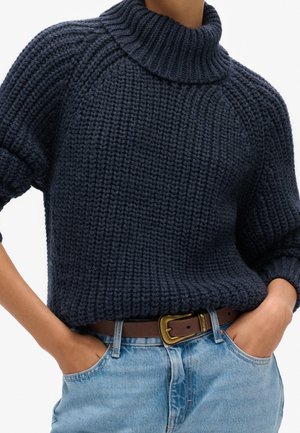 Personne portant un pull à col roulé épais bleu marine en maille côtelée, rentré dans un jean bleu clair avec une ceinture en cuir marron et les mains dans les poches.