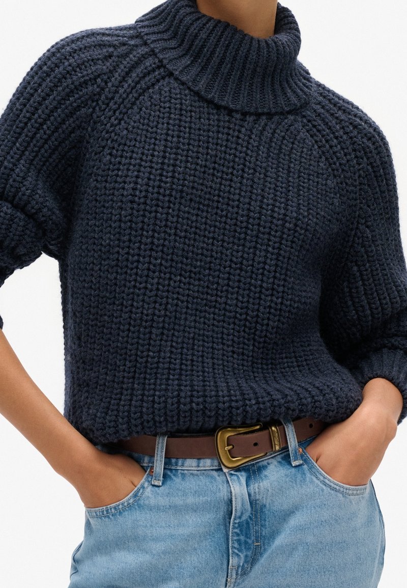 Personne portant un pull à col roulé épais bleu marine en maille côtelée, rentré dans un jean bleu clair avec une ceinture en cuir marron et les mains dans les poches.