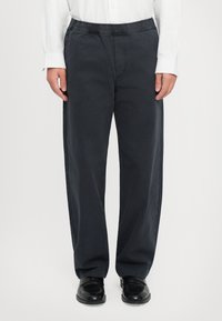 Person wearing loose black pants with elastic waistband, white long-sleeve shirt, and black polished leather shoes, standing on white background.