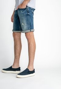 Shorts en denim au lavage foncé avec des détails déchirés et des ourlets roulés, associés à des chaussures à enfiler bleu marine avec une semelle blanche.