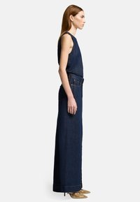 Combinaison en denim bleu foncé. Modèle sans manches, coupe droite avec poches latérales et passants de ceinture. Associée à des talons aiguilles à clous.