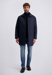 Ein Mann trägt einen marineblauen, geknöpften Mantel, einen dunklen Rollkragenpulli, helle Jeans und dunkle Slipper und steht vor einem schlichten Hintergrund.